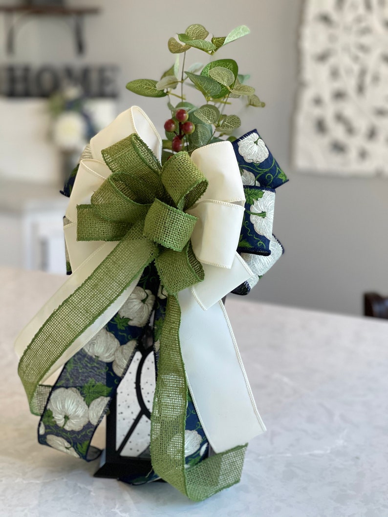K&ouml;nnte beinhalten: Eine dekorative Schleife mit einer Mischung aus Texturen und Farben. Die Schleife enth&auml;lt wei&szlig;en Satin, gr&uuml;nes Jute und marineblaues Band mit wei&szlig;en Blumenmustern. Sie ist mit k&uuml;nstlichem Gr&uuml;n und roten Beeren verziert, geeignet f&uuml;r die Inneneinrichtung.