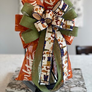 May include: A fall-themed bow with orange, green, and white ribbon featuring a scarecrow and pumpkin pattern. The ribbon is tied around a lantern with the word "thankful" written on it.