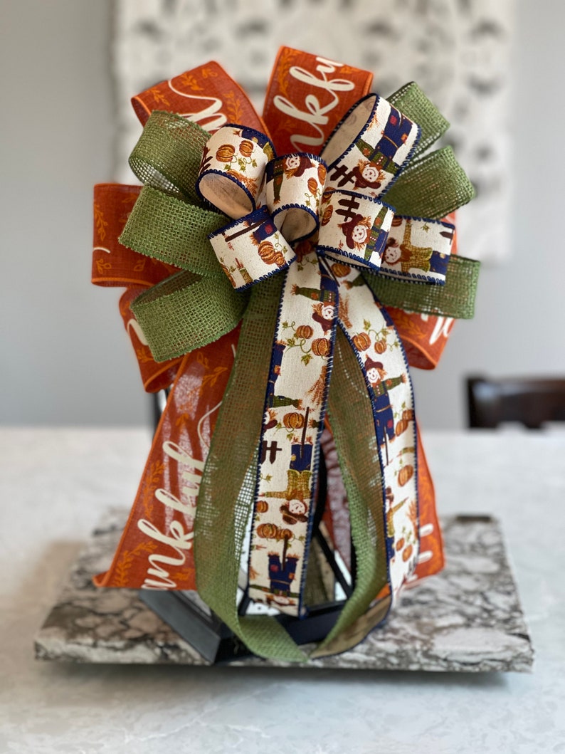 May include: A fall-themed bow with orange, green, and white ribbon featuring a scarecrow and pumpkin pattern. The ribbon is tied around a lantern with the word "thankful" written on it.