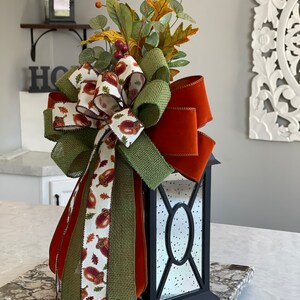 May include: A black lantern with a decorative fall-themed bow and foliage. The bow features a mix of burlap, orange, and acorn-printed ribbons. The lantern is adorned with faux leaves, berries, and a decorative arrangement, perfect for seasonal decor.