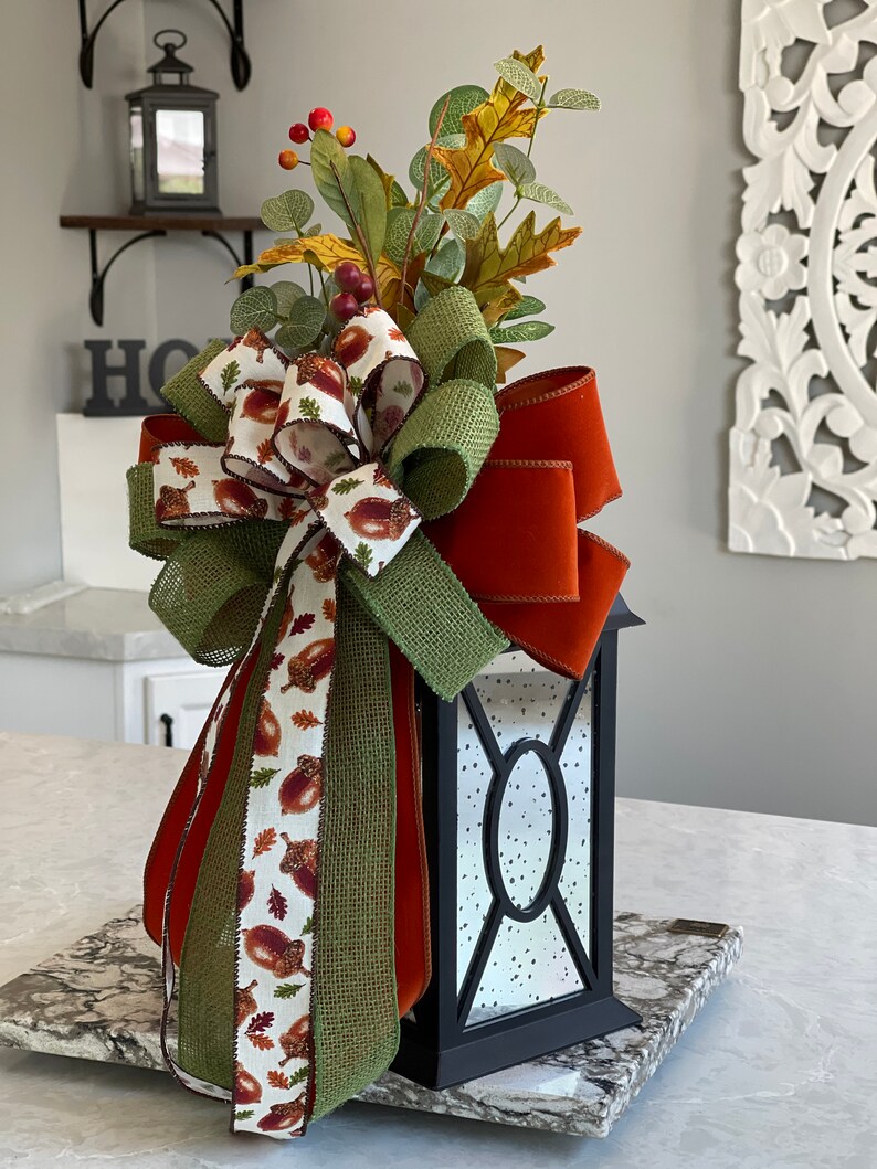 May include: A black lantern with a decorative fall-themed bow and foliage. The bow features a mix of burlap, orange, and acorn-printed ribbons. The lantern is adorned with faux leaves, berries, and a decorative arrangement, perfect for seasonal decor.