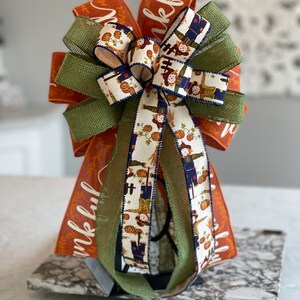 May include: A large, orange, green, and white bow with a burlap ribbon and a pattern of scarecrows and pumpkins. The bow is tied with a black ribbon and the words "Thankful" are printed on the orange ribbon.