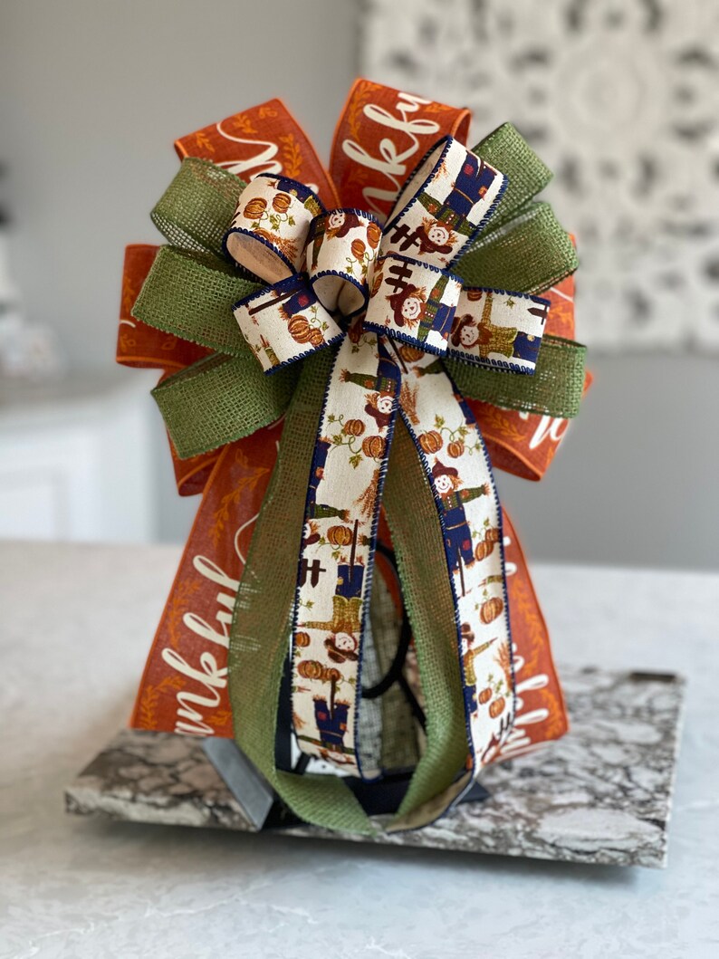May include: A large, orange, green, and white bow with a burlap ribbon and a pattern of scarecrows and pumpkins. The bow is tied with a black ribbon and the words "Thankful" are printed on the orange ribbon.