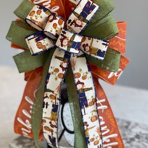 May include: A Thanksgiving-themed bow with orange and green burlap ribbon, a white ribbon with a scarecrow pattern, and the word "thankful" written on the orange ribbon.