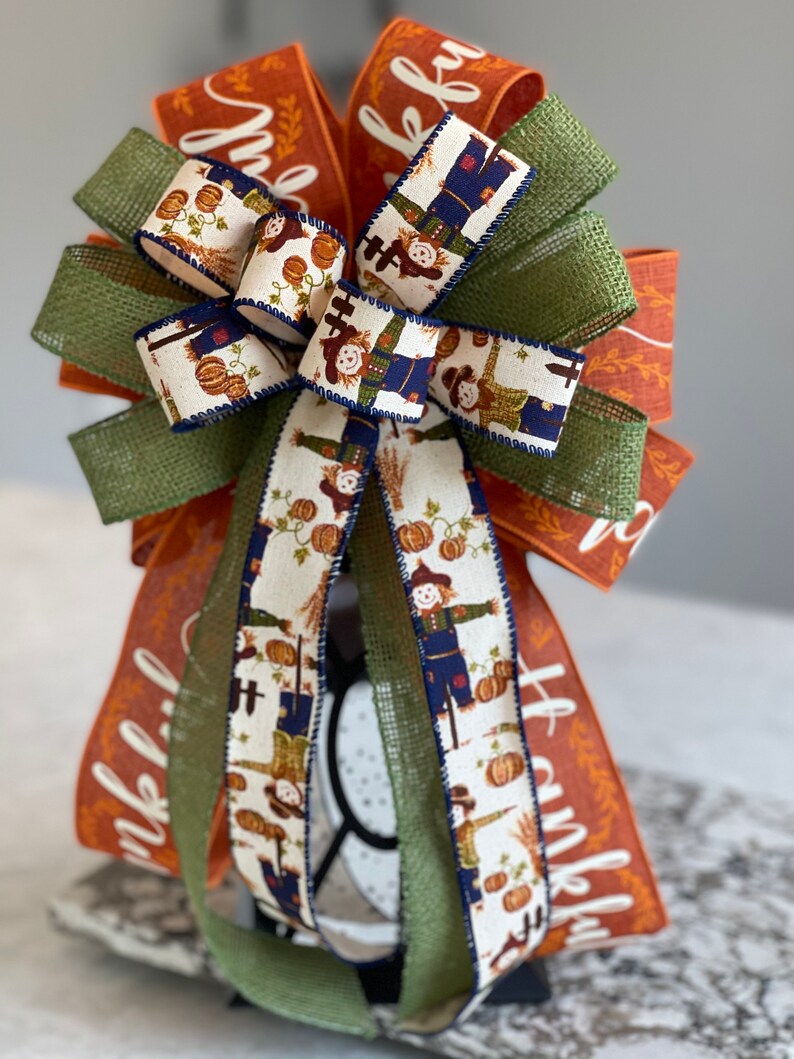 May include: A Thanksgiving-themed bow with orange and green burlap ribbon, a white ribbon with a scarecrow pattern, and the word "thankful" written on the orange ribbon.