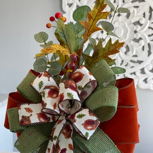 May include: A decorative fall-themed bow with a mix of textures and colors. The bow features a burlap ribbon with a green hue, a solid rust-colored ribbon, and a patterned ribbon with acorns and leaves. It is adorned with faux leaves and berries.