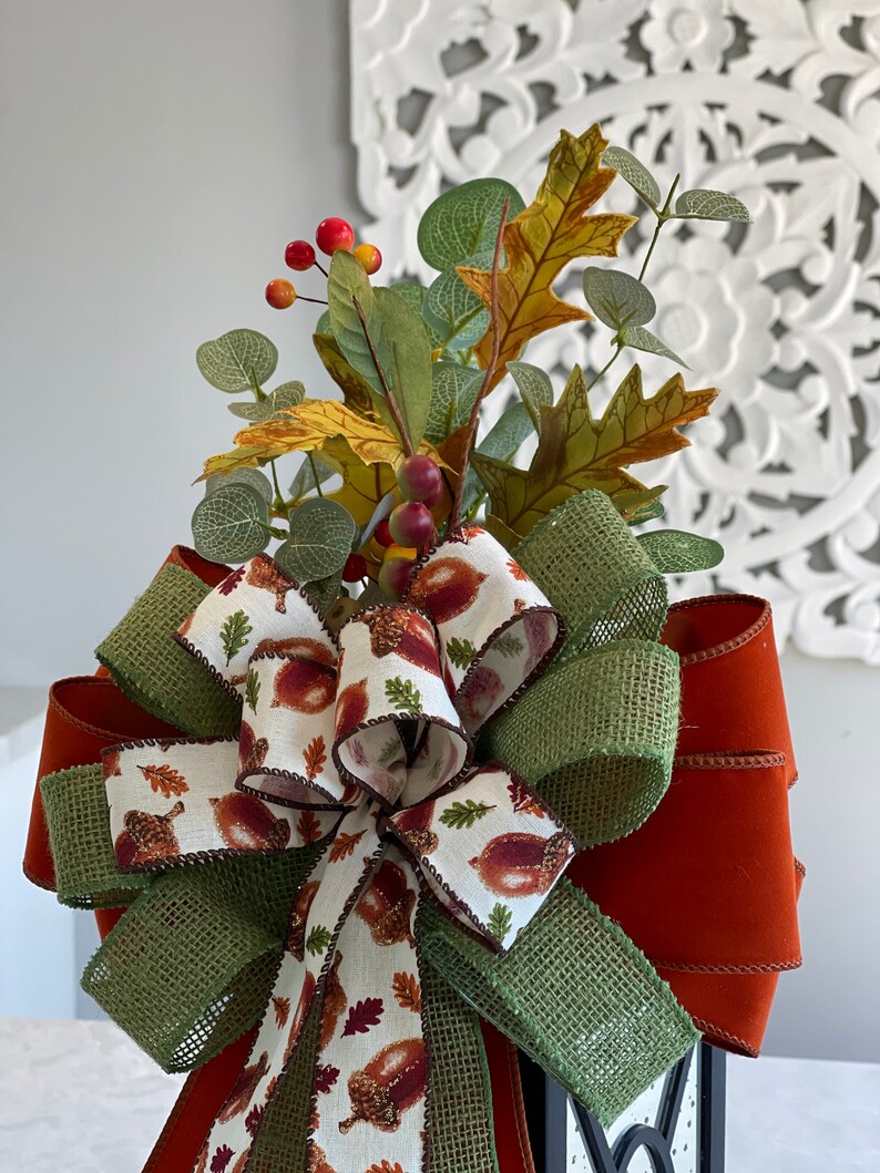 May include: A decorative fall-themed bow with a mix of textures and colors. The bow features a burlap ribbon with a green hue, a solid rust-colored ribbon, and a patterned ribbon with acorns and leaves. It is adorned with faux leaves and berries.