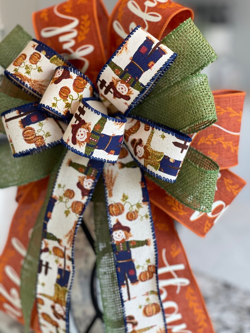 May include: A fall-themed bow with a burlap and green ribbon, and a white ribbon with a pattern of scarecrows and pumpkins. The ribbon has the word "thankful" written on it.
