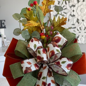 May include: A decorative fall-themed bow featuring a mix of textures and colors. The bow has a large, rust-colored base with layers of green burlap and a white ribbon printed with acorns and leaves. It is topped with faux fall foliage, including leaves and berries.