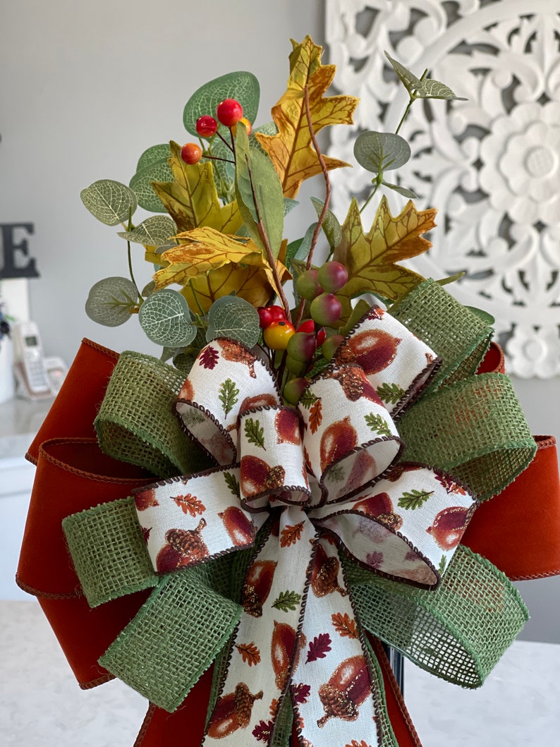 May include: A decorative fall-themed bow featuring a mix of textures and colors. The bow has a large, rust-colored base with layers of green burlap and a white ribbon printed with acorns and leaves. It is topped with faux fall foliage, including leaves and berries.