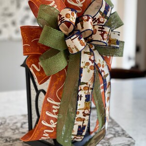 May include: A Thanksgiving-themed bow with orange, green, and white ribbon. The ribbon features the word "thankful" and a pattern of scarecrows and pumpkins. The bow is tied around a black lantern.