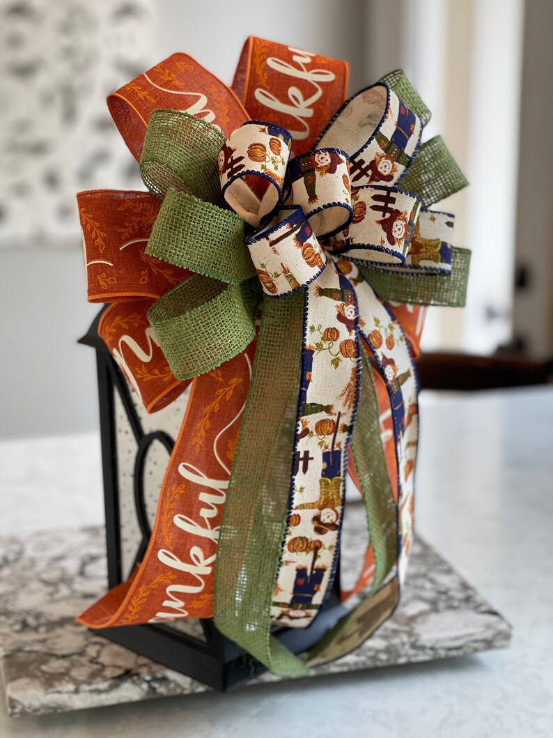 May include: A Thanksgiving-themed bow with orange, green, and white ribbon. The ribbon features the word "thankful" and a pattern of scarecrows and pumpkins. The bow is tied around a black lantern.