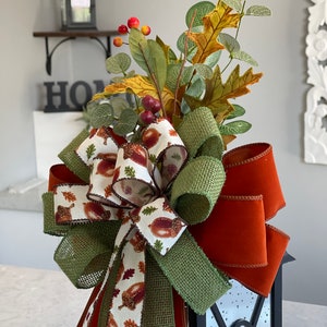 May include: A decorative fall-themed lantern topper featuring a large bow with a burlap and acorn print ribbon, accented by a burnt orange velvet ribbon. The topper is adorned with faux fall foliage, including leaves and berries, creating a festive autumn display.