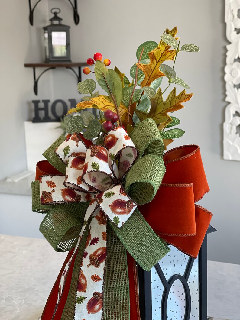 May include: A decorative fall-themed lantern topper featuring a large bow with a burlap and acorn print ribbon, accented by a burnt orange velvet ribbon. The topper is adorned with faux fall foliage, including leaves and berries, creating a festive autumn display.