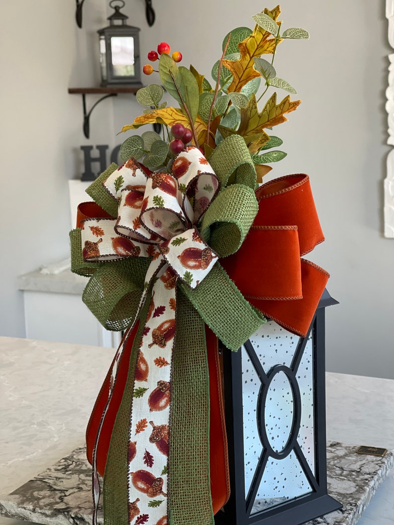 May include: A black lantern decorated with a fall-themed bow and foliage. The bow features a burlap-style green ribbon, a rust-colored ribbon, and a ribbon printed with acorns and leaves. The arrangement includes faux leaves, berries, and greenery.