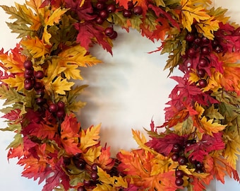 May include: A circular wreath constructed from artificial autumn foliage featuring a mix of serrated maple-style leaves in shades of red, orange, yellow, and green, accented with clusters of small, dark brown berries throughout the arrangement.