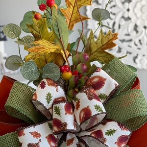 May include: A decorative fall-themed bow featuring a white ribbon printed with acorns and leaves, accented with a green burlap ribbon. The bow is adorned with faux fall foliage, including leaves, berries, and eucalyptus.