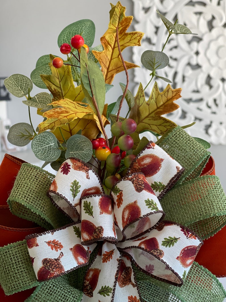 May include: A decorative fall-themed bow featuring a white ribbon printed with acorns and leaves, accented with a green burlap ribbon. The bow is adorned with faux fall foliage, including leaves, berries, and eucalyptus.
