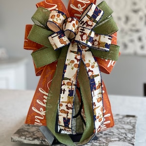 May include: A large, decorative bow made with orange, green, and white ribbon. The ribbon features a pattern of scarecrows and pumpkins. The bow is tied with a green burlap ribbon and has the word "thankful" written on it.