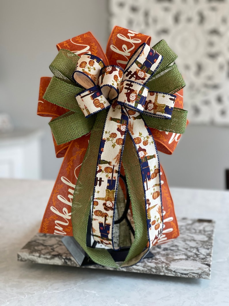 May include: A large, decorative bow made with orange, green, and white ribbon. The ribbon features a pattern of scarecrows and pumpkins. The bow is tied with a green burlap ribbon and has the word "thankful" written on it.