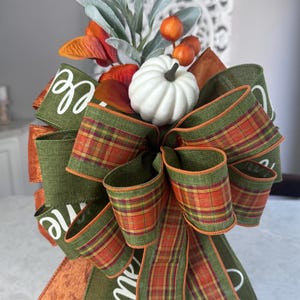May include: A decorative bow featuring olive green ribbon with white text, orange and green plaid ribbon, and a solid orange ribbon. The center is adorned with a small white pumpkin, orange berries, and sage green leaves, creating a seasonal arrangement.