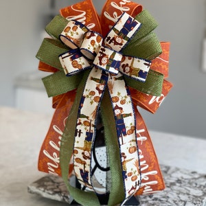 May include: A Thanksgiving-themed bow with orange and green ribbon, featuring a burlap ribbon and a white ribbon with a pattern of scarecrows and pumpkins. The orange ribbon has the word "thankful" written on it.