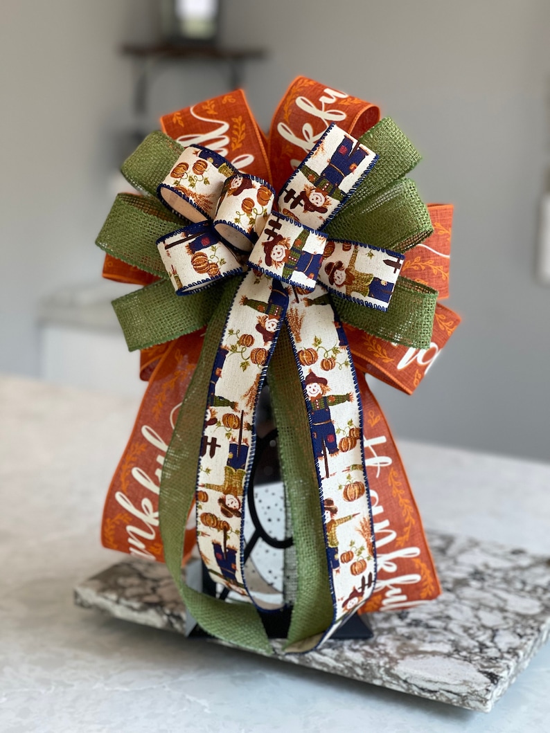 May include: A Thanksgiving-themed bow with orange and green ribbon, featuring a burlap ribbon and a white ribbon with a pattern of scarecrows and pumpkins. The orange ribbon has the word "thankful" written on it.