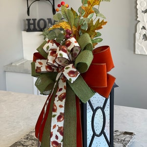May include: A black lantern with a decorative fall-themed bow. The bow features a mix of ribbons in orange, green, and acorn-printed fabric. Artificial leaves and berries top the lantern, creating a festive autumn centerpiece.