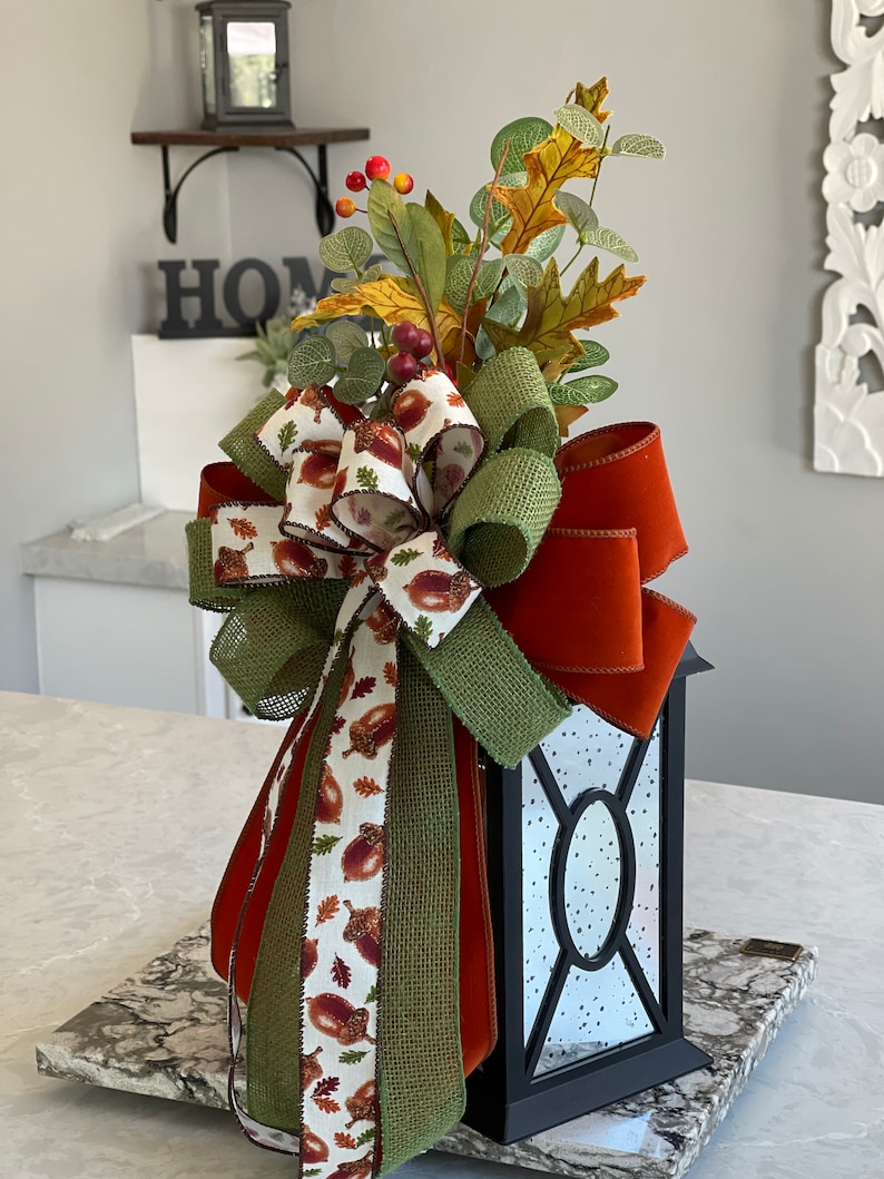 May include: A black lantern with a decorative fall-themed bow. The bow features a mix of ribbons in orange, green, and acorn-printed fabric. Artificial leaves and berries top the lantern, creating a festive autumn centerpiece.