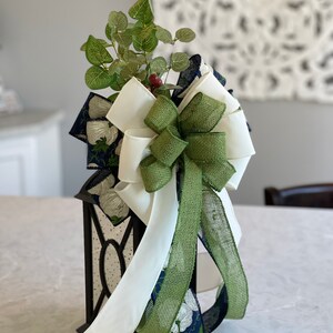 K&ouml;nnte beinhalten: Eine schwarze Laterne mit einer dekorativen Schleife und Gr&uuml;n. Die Schleife hat wei&szlig;e, gr&uuml;ne und marineblaue B&auml;nder mit einem floralen Muster. Die Laterne hat transparente Paneele und ist mit k&uuml;nstlichem Gr&uuml;n und roten Beeren verziert.
