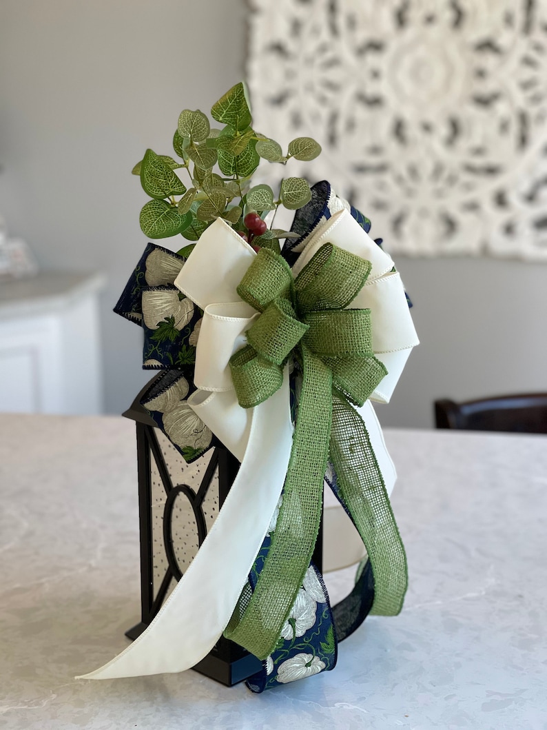 K&ouml;nnte beinhalten: Eine schwarze Laterne mit einer dekorativen Schleife und Gr&uuml;n. Die Schleife hat wei&szlig;e, gr&uuml;ne und marineblaue B&auml;nder mit einem floralen Muster. Die Laterne hat transparente Paneele und ist mit k&uuml;nstlichem Gr&uuml;n und roten Beeren verziert.