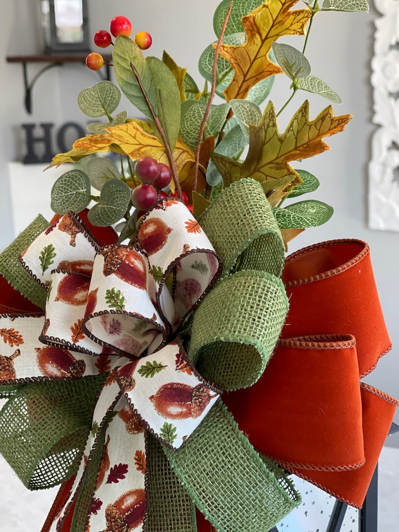 May include: A close-up of a decorative fall-themed bow. The bow features a mix of textures and colors, including orange, green, and cream ribbons. The cream ribbon is printed with acorns. The bow is adorned with faux leaves and berries.