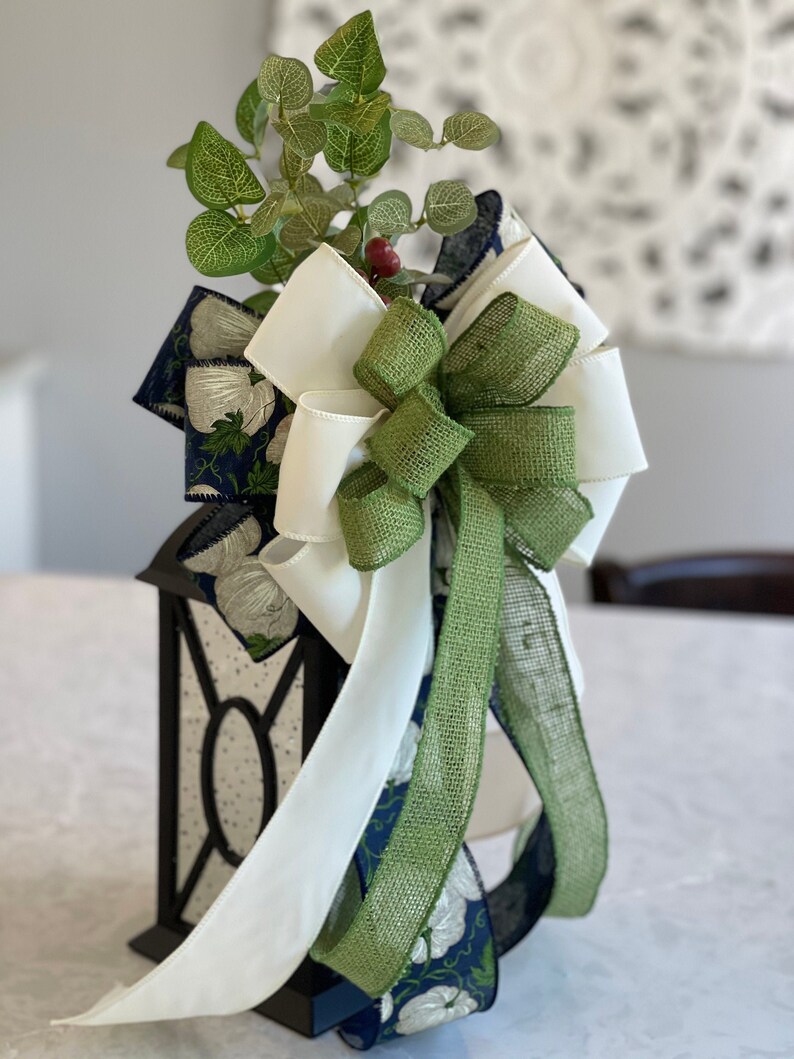 K&ouml;nnte beinhalten: Eine schwarze Laterne mit einer wei&szlig;en und gr&uuml;nen Schleife und Gr&uuml;nzeug. Die Schleife hat wei&szlig;e, gr&uuml;ne und marineblaue B&auml;nder. Das marineblaue Band hat ein wei&szlig;es Blumenmuster. Die Laterne hat ein dekoratives Glasdesign.