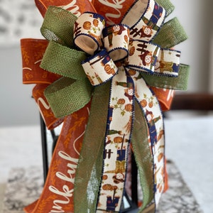 May include: A festive fall-themed bow with orange, green, and white ribbon. The ribbon features a pattern of scarecrows and pumpkins. The bow is tied around a black lantern with the word "thankful" printed on the orange ribbon.