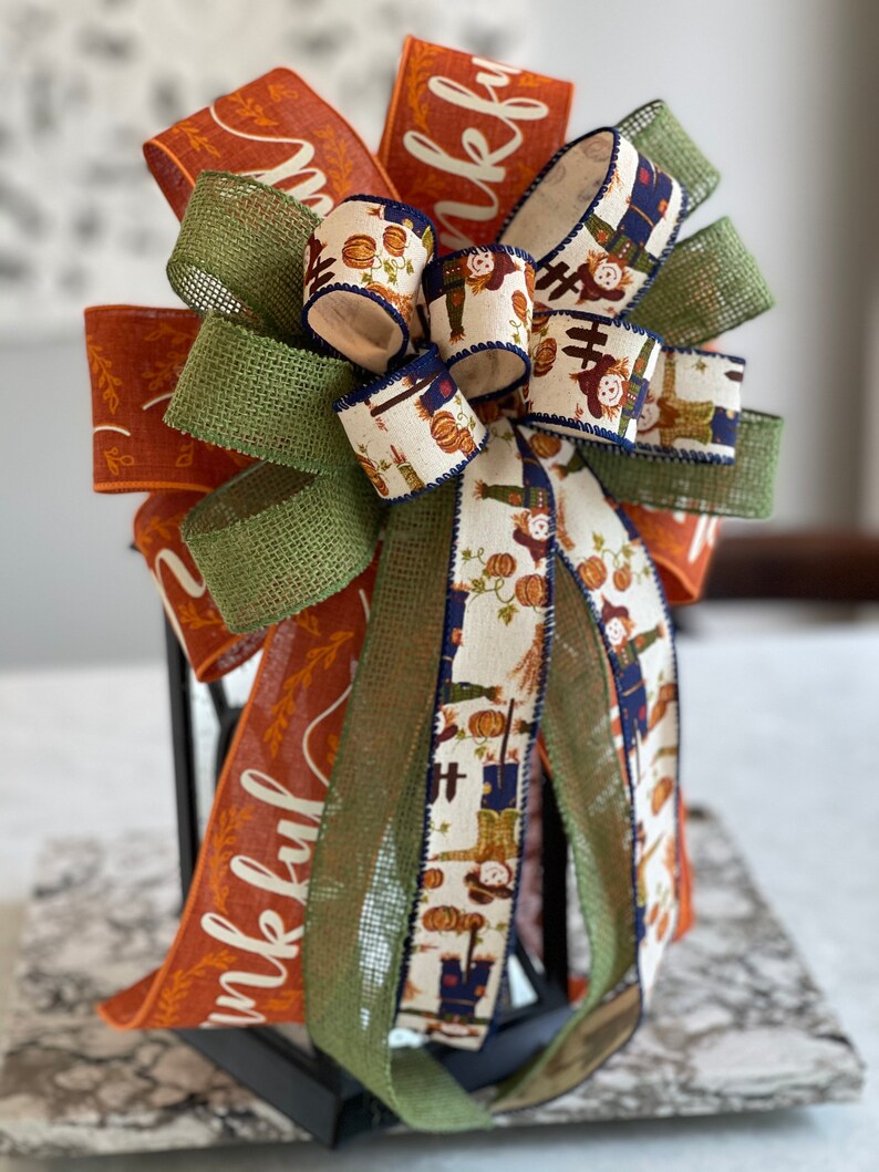 May include: A festive fall-themed bow with orange, green, and white ribbon. The ribbon features a pattern of scarecrows and pumpkins. The bow is tied around a black lantern with the word "thankful" printed on the orange ribbon.