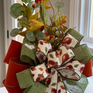 May include: A fall-themed decorative bow featuring orange, green, and cream ribbons. The bow is adorned with faux leaves, berries, and eucalyptus. The cream ribbon has a pattern of acorns and leaves, perfect for autumn decor.