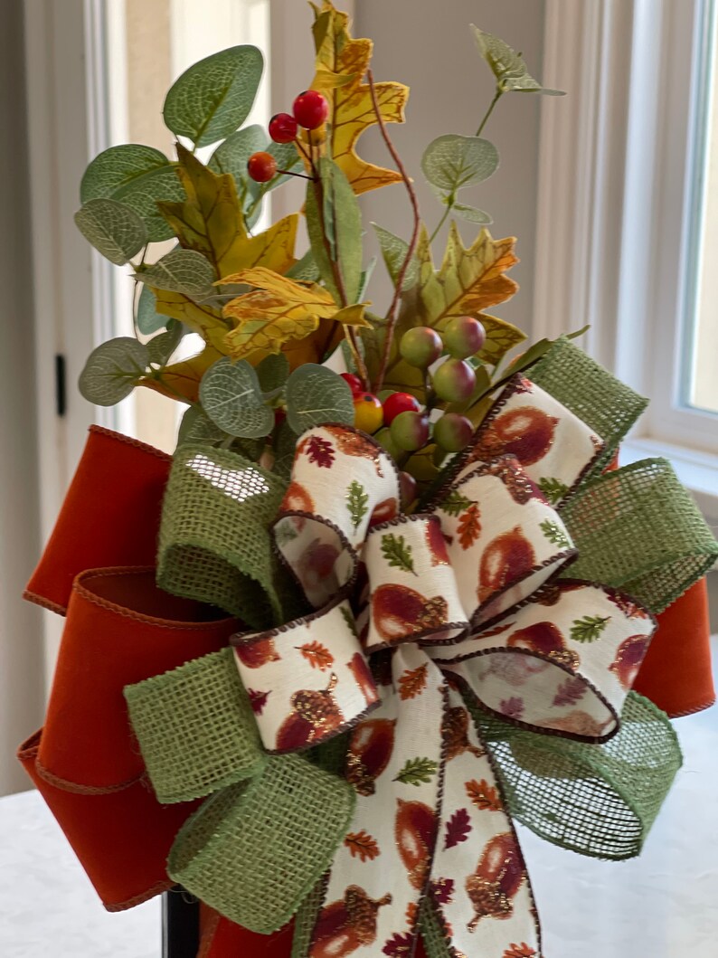 May include: A fall-themed decorative bow featuring orange, green, and cream ribbons. The bow is adorned with faux leaves, berries, and eucalyptus. The cream ribbon has a pattern of acorns and leaves, perfect for autumn decor.