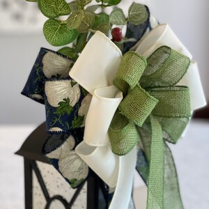 K&ouml;nnte beinhalten: Eine schwarze Laterne, verziert mit einer gro&szlig;en Schleife aus wei&szlig;en, gr&uuml;nen und marineblauen B&auml;ndern. Das marineblaue Band zeigt ein wei&szlig;es Blumenmuster. Gr&uuml;ne Bl&auml;tter und eine kleine rote Beere erg&auml;nzen das Arrangement.