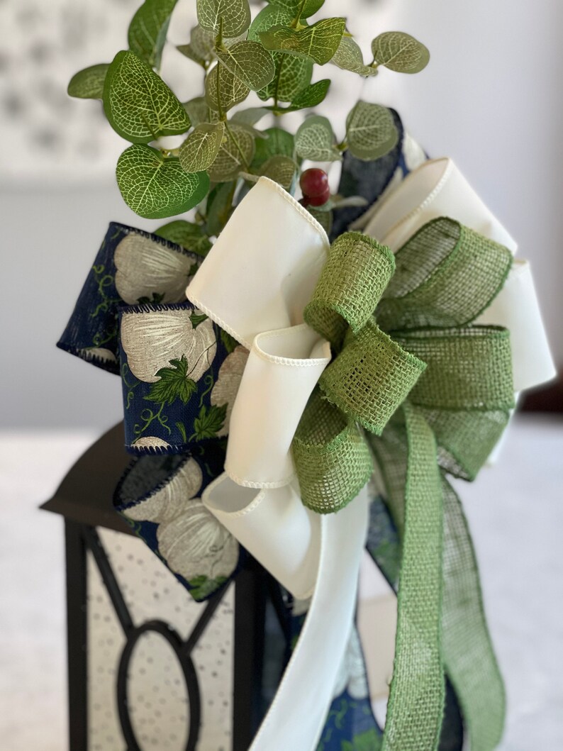 K&ouml;nnte beinhalten: Eine schwarze Laterne, verziert mit einer gro&szlig;en Schleife aus wei&szlig;en, gr&uuml;nen und marineblauen B&auml;ndern. Das marineblaue Band zeigt ein wei&szlig;es Blumenmuster. Gr&uuml;ne Bl&auml;tter und eine kleine rote Beere erg&auml;nzen das Arrangement.