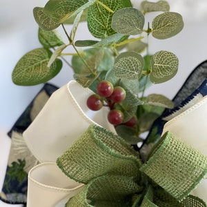 K&ouml;nnte beinhalten: Nahaufnahme einer dekorativen Schleife mit einer gr&uuml;nen Jute-Schleife, wei&szlig;em Satinband und marineblauem, gemustertem Band. Eukalyptusbl&auml;tter und rote Beeren sind ebenfalls enthalten. Geeignet f&uuml;r Bastelprojekte und Dekorationen.
