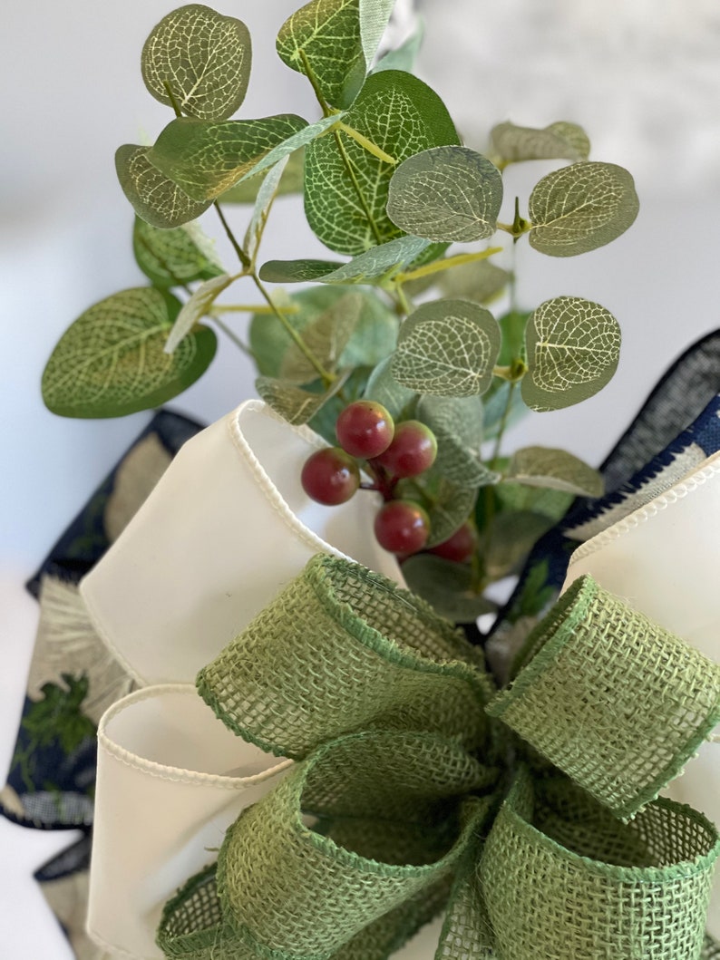 K&ouml;nnte beinhalten: Nahaufnahme einer dekorativen Schleife mit einer gr&uuml;nen Jute-Schleife, wei&szlig;em Satinband und marineblauem, gemustertem Band. Eukalyptusbl&auml;tter und rote Beeren sind ebenfalls enthalten. Geeignet f&uuml;r Bastelprojekte und Dekorationen.