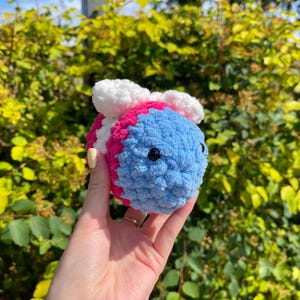 May include: A crocheted bee toy in blue, pink, and white. The bee has black eyes and white wings. The plush toy is held in a hand against a backdrop of green leaves.