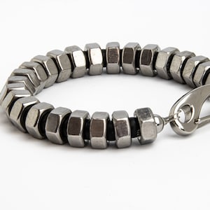 Könnte beinhalten: Ein silberfarbenes Armband aus sechseckigen Metallmuttern, die miteinander verbunden sind. Das Armband hat einen silbernen Verschluss und einen kleinen, runden Anhänger. Das industrielle Design verleiht dem Armband eine einzigartige, kantige Ästhetik.