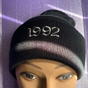 May include: Black knitted beanie with a white embroidered "1992" on the front.