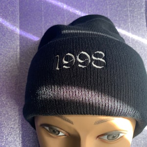 Könnte beinhalten: Schwarze Strickmütze mit einem weißen gestickten "1998" auf der Vorderseite.