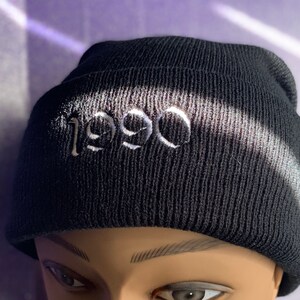 May include: Black knitted beanie hat with the year "1990" embroidered in white. The hat is made of a ribbed knit material and is shown on a mannequin head. The background is a light purple colour.