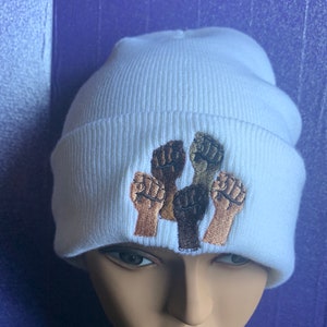 May include: White beanie with an embroidered design of six raised fists in various skin tones. The beanie is made of a soft, ribbed knit material. The fists are in shades of brown, tan, and black.