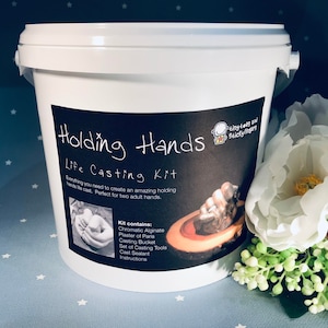 May include: A white bucket labelled "Holding Hands Life Casting Kit." The kit contains materials to create a holding hands life cast. The label features an illustration of hands clasped together. The kit is for two adult hands.