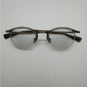 May include: Round-framed eyeglasses with a brown metal frame and tinted lenses. The temples have a tortoiseshell pattern. The glasses are set against a white background.