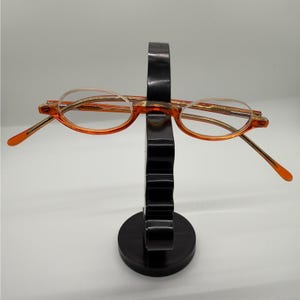 Puede incluir: Gafas con tinte naranja y montura dorada sobre un expositor negro. Las lentes ovaladas son claras, y las patillas naranjas. El expositor negro tiene una base circular, ideal para gafas.